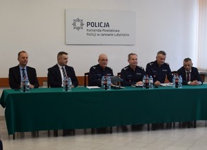odprawa roczna przy stole siedzą osoby biorące udział w odprawie od lewej Burmistrz Janowa Lubelskiego Krzysztof Kołtyś, obok Starosta Janowski Artur Pizoń, kolejno Komendant Powiatowy Policji w Janowie Lubelskim podinspektor Stanisław Niemczycki następnie Zastępca Komendanta Wojewódzkiego Policji w Lublinie inspektor Andrzej Mioduna, następnie I Zastępca Komendanta Powiatowego Policji w Janowie Lubelskim młodszy inspektor Stanisław Tyburski następnie Prokurator Rejonowy w Janowie Lubelskim Radosław Lisek